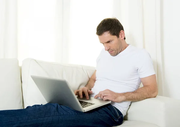 Красивый привлекательный мужчина дома лежит на диване и работает фрилансером с ноутбуком — стоковое фото