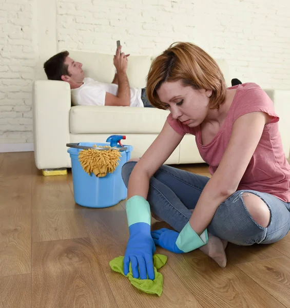 Муж отдыхает на диване, в то время как жена убирает, выполняя работу по дому в концепции шовинизма — стоковое фото
