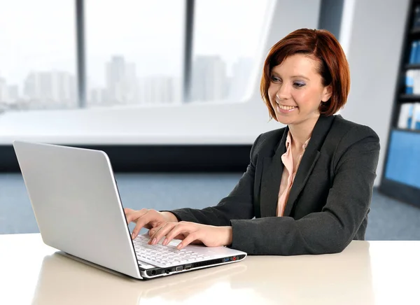 Счастливая деловая женщина с рыжими волосами, улыбающаяся на работе, печатающая на ноутбуке в современном офисе — стоковое фото