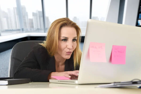 Расслабленная 40-летняя женщина с светлыми волосами, уверенно улыбающаяся, сидя на офисном стуле и работая за ноутбуком — стоковое фото