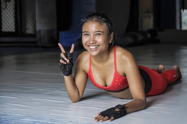 Young happy and beautiful sweaty Asian woman in sport clothes stretching on gym dojo hy sporty lifestyle concept giving peace sign — Stock Photo, Image