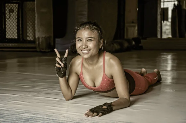 Young happy and beautiful sweaty Asian woman in sport clothes stretching on gym dojo hy sporty lifestyle concept giving peace sign — Stock Photo, Image