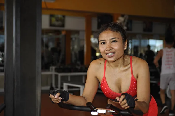 Hermosa sudorosa asiática feliz mujer entrenamiento sonriendo duro ciclismo y montar en bicicleta estática entrenamiento en el gimnasio — Foto de Stock