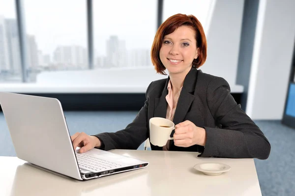 Business woman with red hair at work smiling on laptop computer desk and drinking coffee — Stock Photo, Image