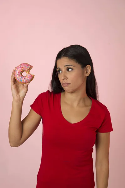 Mooie Latijns jongedame in rood met roze suiker donut denken en schuldgevoel na bijten geïsoleerd — Stockfoto