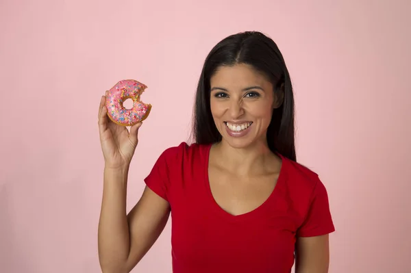 Молодая счастливая привлекательная латиноамериканка в красном топе улыбается взволнованный держа сахарный пончик изолирован на розовом фоне — стоковое фото