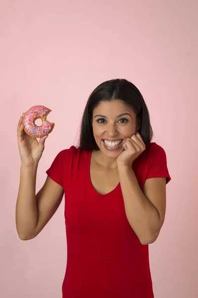 Mladá šťastná atraktivní Latin žena v červený top s úsměvem nadšený, drží cukru kobliha izolované na růžovém pozadí — Stock fotografie