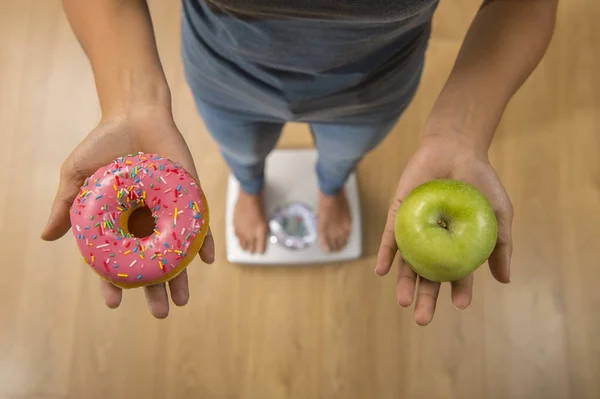 Close up woman on weight scale holding in her hand apple fruit and donut as choice of healthy versus unhealthy food — Stock Photo, Image