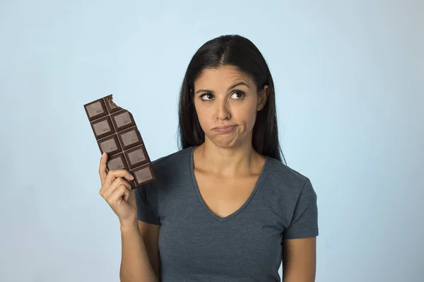 Bella donna latina con barretta di cioccolato sentirsi in colpa dopo aver morso isolato su sfondo blu in zucchero e dolce abuso — Foto Stock