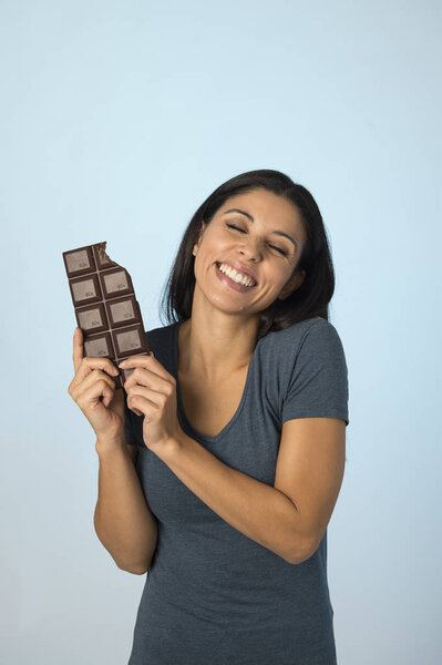 young attractive and happy hispanic woman in blue top smiling excited eating chocolate bar isolated background
