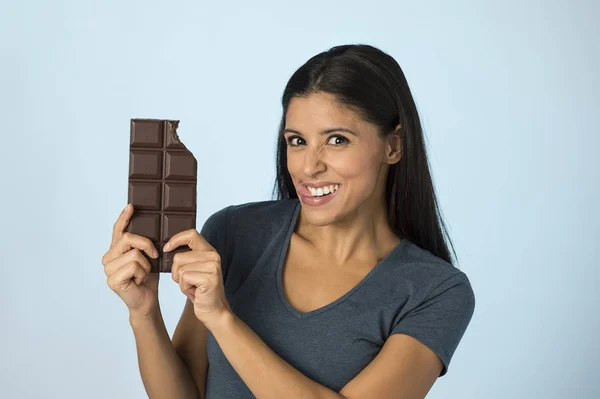Aantrekkelijk en gelukkig hispanic jongedame in blauwe top glimlachend opgewonden eten chocolade bar geïsoleerde achtergrond — Stockfoto