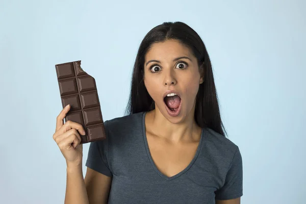 Aantrekkelijk en gelukkig hispanic jongedame in blauwe top glimlachend opgewonden eten chocolade bar geïsoleerde achtergrond — Stockfoto