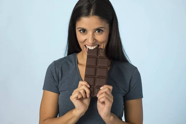 Young attractive and happy hispanic woman in blue top smiling excited eating chocolate bar isolated background — Stock Photo, Image