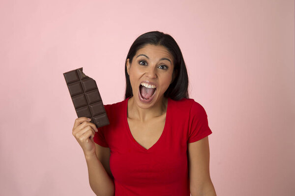 young attractive and happy hispanic woman in red top smiling excited eating chocolate bar isolated on pink background