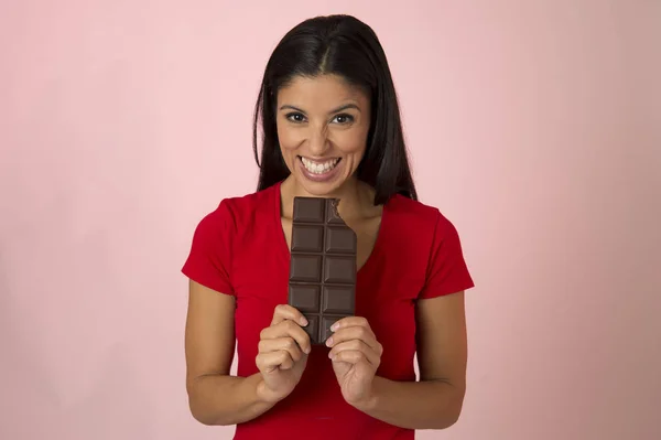 Giovane donna ispanica attraente e felice in top rosso sorridente eccitato mangiare cioccolato bar isolato su sfondo rosa — Foto Stock