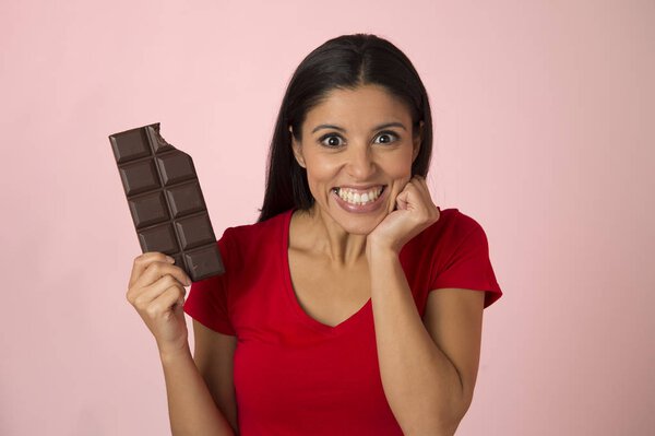 young attractive and happy hispanic woman in red top smiling excited eating chocolate bar isolated on pink background