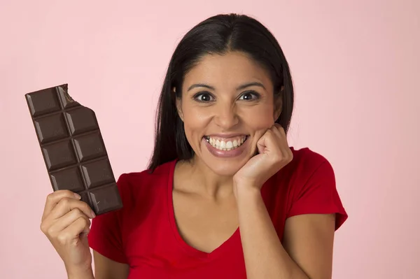 Junge attraktive und glückliche hispanische Frau in rotem Top lächelnd aufgeregt essen Schokoriegel isoliert auf rosa Hintergrund — Stockfoto