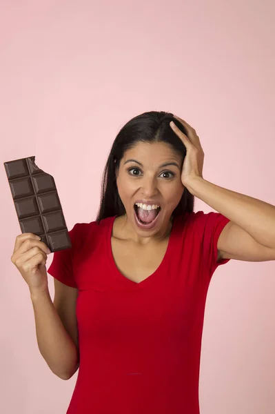 Junge attraktive und glückliche hispanische Frau in rotem Top lächelnd aufgeregt essen Schokoriegel isoliert auf rosa Hintergrund — Stockfoto