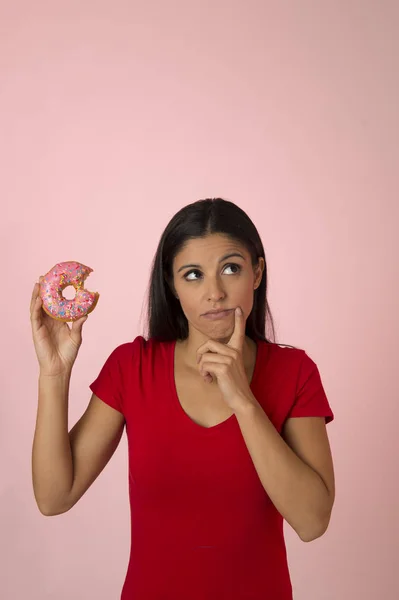 Ung vacker latin kvinna i rött med rosa sockret donut tänka och känna skuld efter bita isolerade — Stockfoto