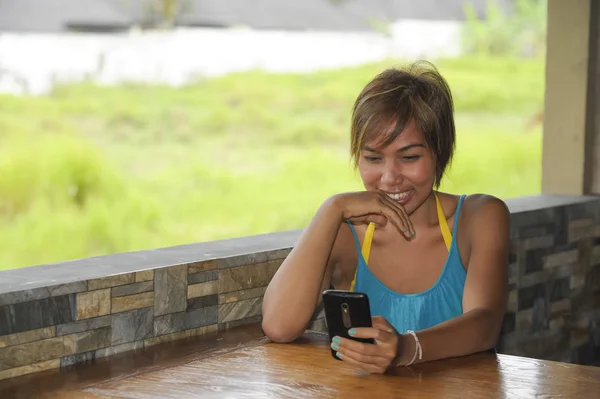 Młody szczęśliwy piękne i atrakcyjne Asian kobieta siedząc w kawiarni przy użyciu aplikacji internet na telefon komórkowy, śmiejąc się podekscytowany na zewnątrz — Zdjęcie stockowe