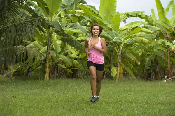 Junge attraktive asiatische Sportlerin, die im Dschungel läuft, lächelt glücklich beim Training auf Kräutern mit Palmen im Hintergrund — Stockfoto