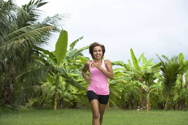 Junge attraktive asiatische Sportlerin, die im Dschungel läuft, lächelt glücklich beim Training auf Kräutern mit Palmen im Hintergrund — Stockfoto