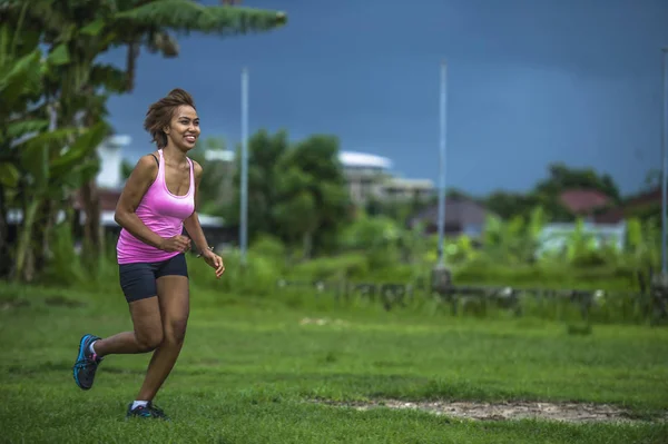 Junge attraktive asiatische Sportlerin, die im Dschungel läuft, lächelt glücklich beim Training auf Kräutern mit Palmen im Hintergrund — Stockfoto