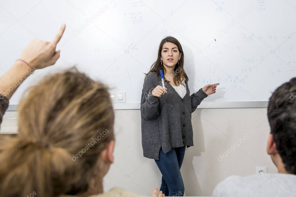 group of young students taking lesson at University classroom with girl standing on blackboard explaining and pupils sitting on desk 