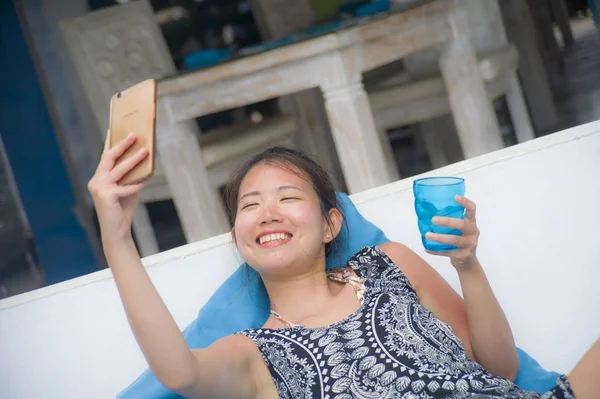 Young sweet and pretty Asian Chinese woman sitting at beach holiday resort taking selfie picture on mobile phone happy and excited — Stock Photo, Image