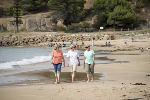 Kelompok yang terdiri dari tiga perempuan dewasa berusia 60-an yang menikmati berjalan-jalan dengan gembira di pantai dengan tersenyum gembira — Stok Foto