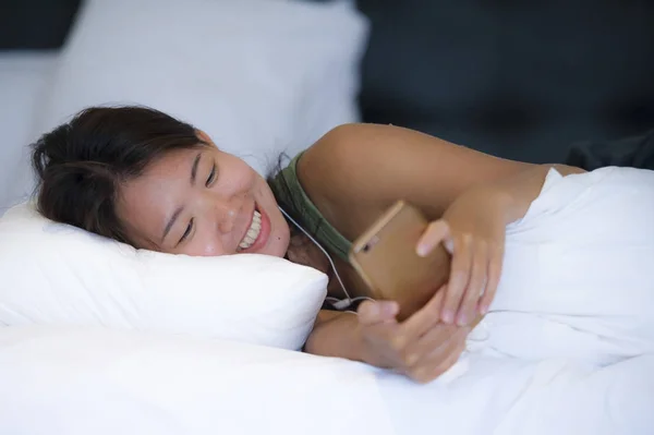 Молодая красивая и расслабленная азиатская китаянка улыбается счастливо лежа на кровати в спальне с помощью интернета на мобильном телефоне — стоковое фото