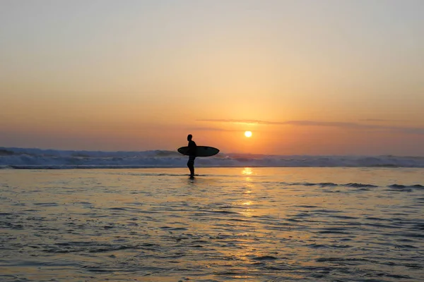 Silueta de surfista anónimo desconocido sosteniendo tabla de surf después de surfear en la puesta del sol con increíble luz solar hermosa con cielo naranja — Foto de Stock
