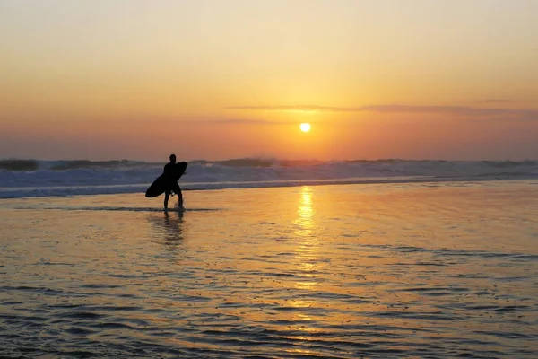 Silhouette of unknown anonymous surfer holding surf board after surfing on sunset with amazing beautiful sunlight with orange sky — Stock Photo, Image