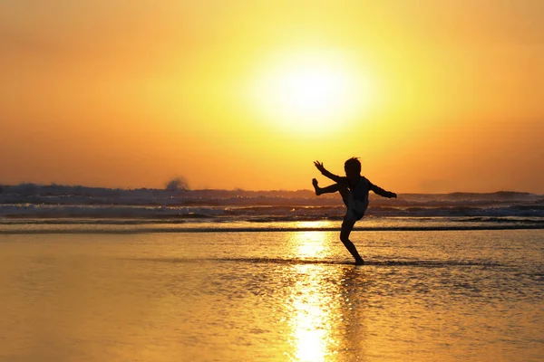 Silhouette of anonymous unknown young kid having fun playing on sea water at the beach kicking on wet sand with amazing beautiful sunset — Stock Photo, Image