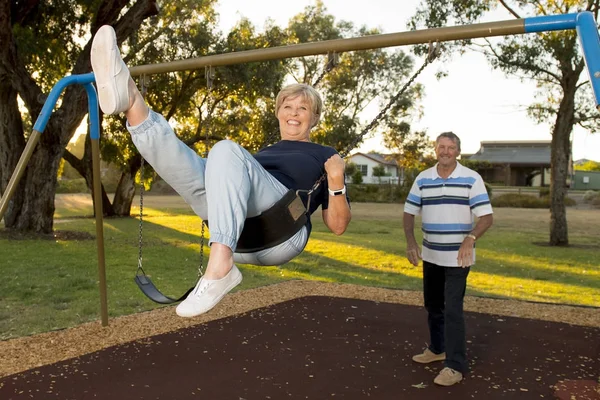 70岁左右的快乐的美国夫妇在摇摆公园享受与丈夫推妻子微笑和有乐趣 — 图库照片