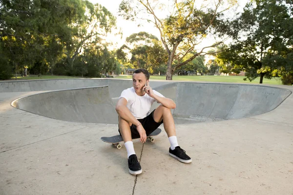 Junger glücklicher und attraktiver amerikanischer Mann in den 30er Jahren sitzt nach dem Sportinternat auf dem Skateboard und telefoniert mit dem Handy — Stockfoto