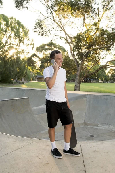 Jovem feliz e atraente americano 30 anos em pé segurando skate board após esporte embarque sessão de treinamento falando no telefone móvel — Fotografia de Stock