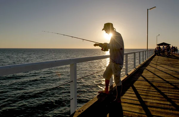 Siluet nelayan dengan topi dan batang ikan berdiri di dermaga laut memancing saat matahari terbenam dengan langit oranye yang indah dalam liburan santai hobi — Stok Foto