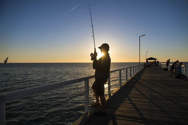 休暇で美しいオレンジ色の空と夕日に海のドック釣りに立っている帽子と魚の棒と漁師のシルエットは、趣味をリラックス — ストック写真