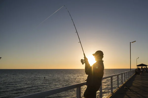 Silhouette eines Fischers mit Hut und Angelrute, der auf dem Dock steht, Angeln bei Sonnenuntergang mit wunderschönem orangefarbenem Himmel im Urlaub entspannen Hobby — Stockfoto