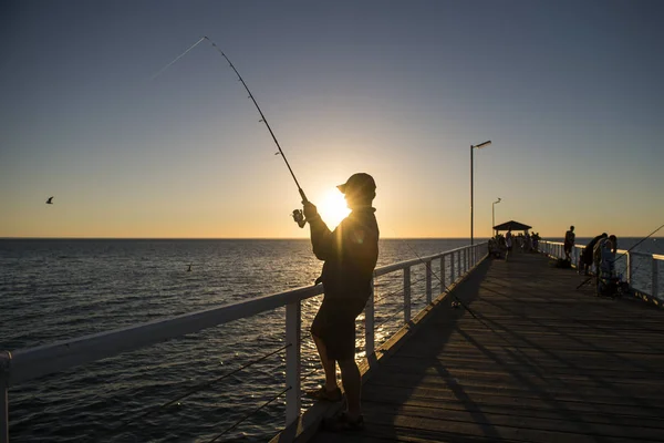 Siluet nelayan dengan topi dan batang ikan berdiri di dermaga laut memancing saat matahari terbenam dengan langit oranye yang indah dalam liburan santai hobi — Stok Foto