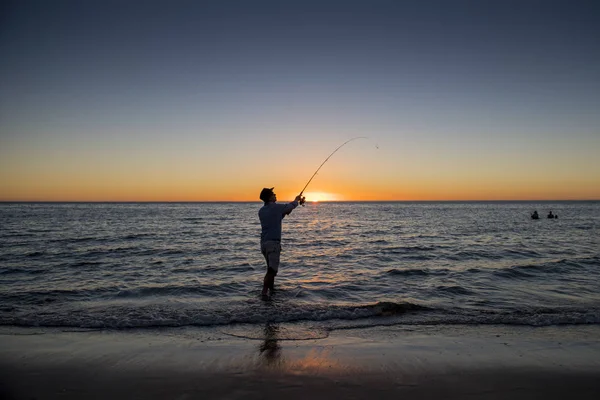 休暇で美しいオレンジ色の空と夕日に海水釣りに立っている魚の棒とビーチで帽子を持つ漁師のシルエット — ストック写真