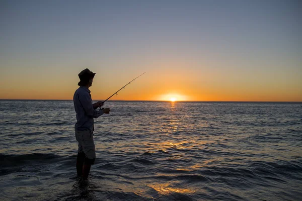 Siluet nelayan dengan topi di pantai dengan batang ikan berdiri di atas air laut memancing saat matahari terbenam dengan langit oranye yang indah dalam liburan — Stok Foto