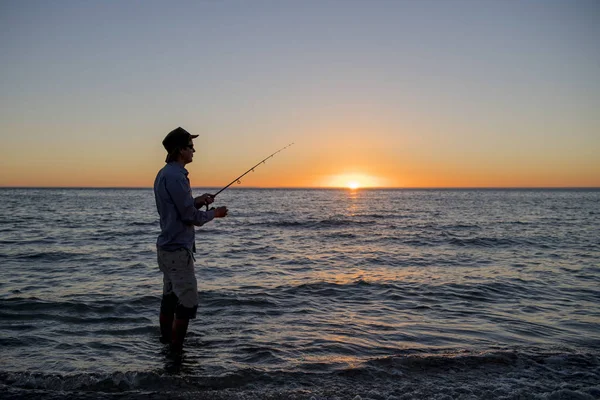 Siluet nelayan dengan topi di pantai dengan batang ikan berdiri di atas air laut memancing saat matahari terbenam dengan langit oranye yang indah dalam liburan — Stok Foto