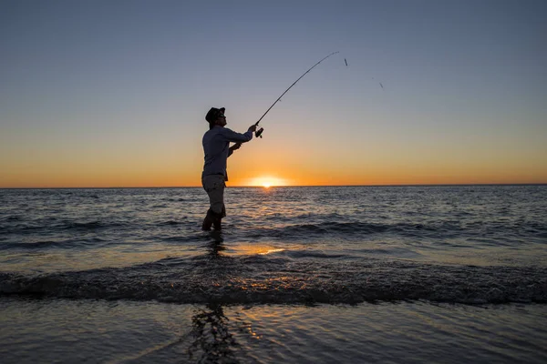 Siluet nelayan dengan topi di pantai dengan batang ikan berdiri di atas air laut memancing saat matahari terbenam dengan langit oranye yang indah dalam liburan — Stok Foto