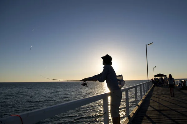 Silueta de pescador con sombrero y caña de pescar de pie en el muelle del mar pesca al atardecer con hermoso cielo naranja en vacaciones relajarse hobby — Foto de Stock