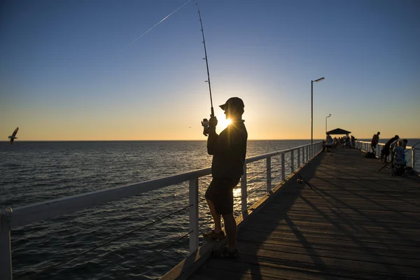 Силуэт рыбака в шляпе и удочке, стоящий на морском причале рыбалки на закате с красивым оранжевым небом в отпуске расслабиться хобби Стоковая Картинка