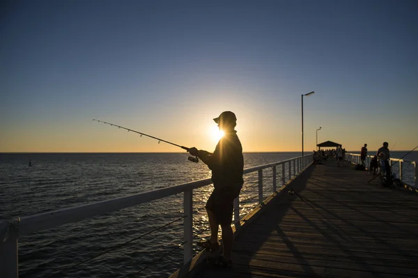 渔夫与帽子和鱼竿站在海上码头钓鱼在日落与美丽的橙色天空在假期放松爱好剪影 图库图片
