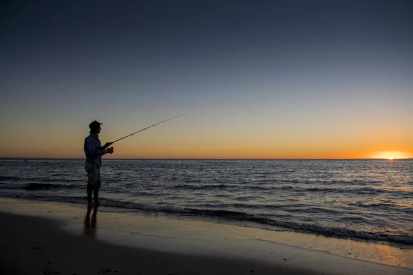 Силуэт рыбака в шляпе на пляже с удочкой, стоящей на морской воде рыбалки на закате с красивым оранжевым небом в отпуске Стоковая Картинка
