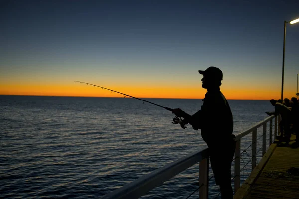 Silhouet van visser met hoed en vis Rod staande op zee dok vissen bij zonsondergang met prachtige oranje hemel in vakantie ontspannen hobby Rechtenvrije Stockafbeeldingen